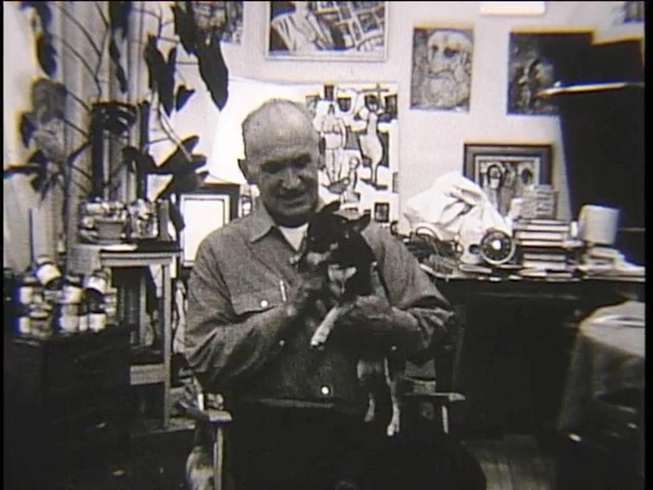 Frank Koci in his studio holding a small dog, surrounded by artwork and creative materials